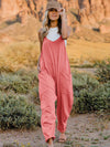 Woman wearing the Double Take Full Size Sleeveless V-Neck Pocketed Jumpsuit in coral pink, styled casually with sandals and a hat in a scenic outdoor setting.

