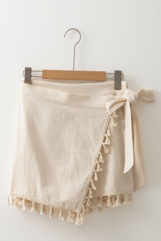 Neutral beige tassel-trimmed wrap skirt shown from the front, displayed flat with tie waist—lightweight fabric ideal for summer and holiday outfits.