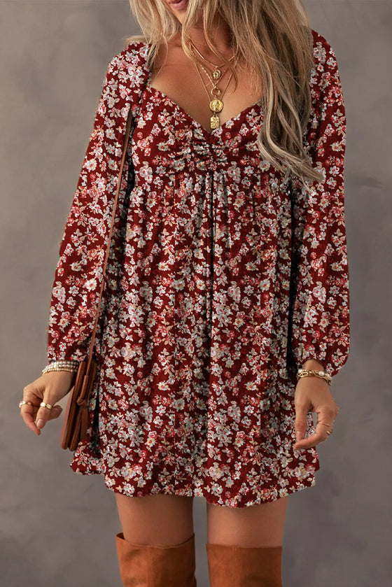 Woman wearing a Ruched Printed Long Sleeve Mini Dress in a floral design, featuring a ruched bodice and mini hemline, styled with boots and layered jewelry.

