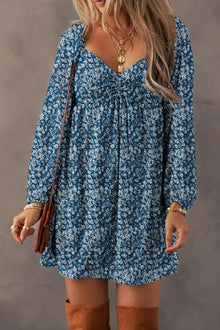  Woman wearing a Ruched Printed Long Sleeve Mini Dress in a floral design, featuring a ruched bodice and mini hemline, styled with boots and layered jewelry.

