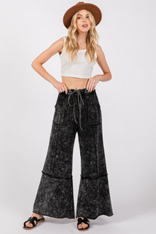  Model wearing black SAGE + FIG Mineral Washed Terry Wide Leg Pants featuring exposed seams, side pockets, and a drawstring waistband, styled with a cropped tank top and sandals.

