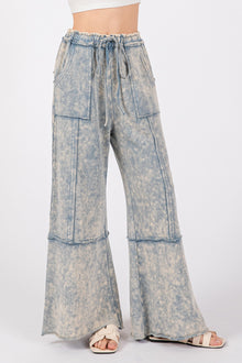  Close-up view of blue SAGE + FIG Mineral Washed Terry Wide Leg Pants with exposed seams, side pockets, and an elastic waistband, styled with sandals and a crop top.

