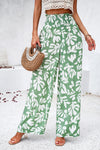 Woman wearing Smocked Printed Wide Leg Pants in green with a bold print, featuring a smocked waistband and side pockets.