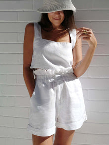  Woman wearing a white square neck sleeveless top with matching high-waist shorts, featuring a ruffled waistband and side pockets. Perfect for casual summer wear.

