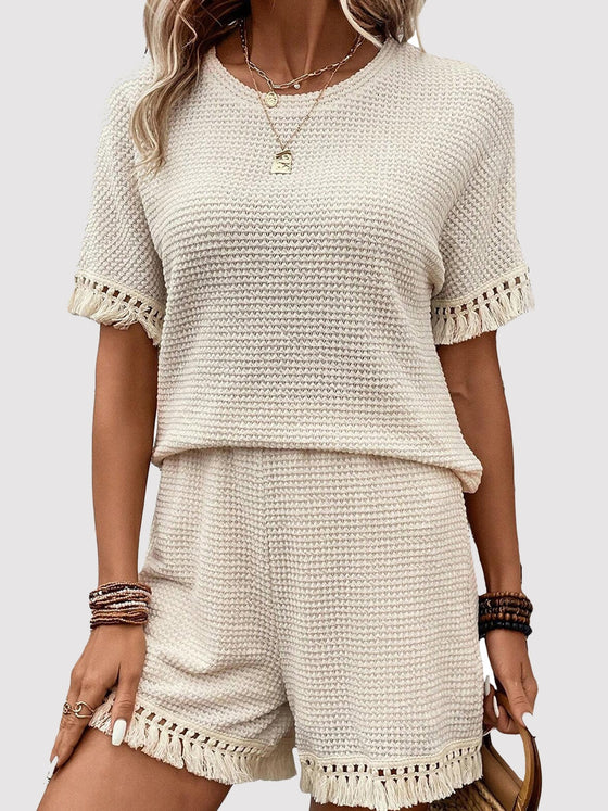 Model wearing a tassel round neck top and shorts set in a beige knit fabric, featuring tassel accents on the sleeves and hem for a boho-inspired look.