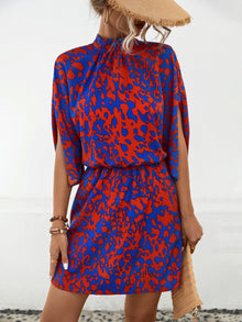  Woman wearing a vibrant Tied Printed Mock Neck Mini Dress in bold red and blue patterns, featuring a mock neck, elastic waist, and slit sleeves.

