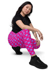 Sitting view of a woman while wearing Vibrant Neon Heart violet-red high-waisted leggings with pockets. Perfect for yoga, gym, or casual wear. High-rise compression leggings with a sleek design and bold neon heart print.