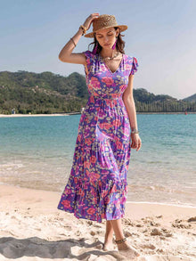  A woman standing on the beach wearing a vibrant floral Printed V-Neck Midi Dress with flutter sleeves and a ruffled A-line hem, styled with a straw hat and jewelry.