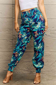  Woman wearing smocked waistband long pants with a tropical plant print, featuring vibrant green leaves and flowers on a navy blue background, paired with a white top and heels.